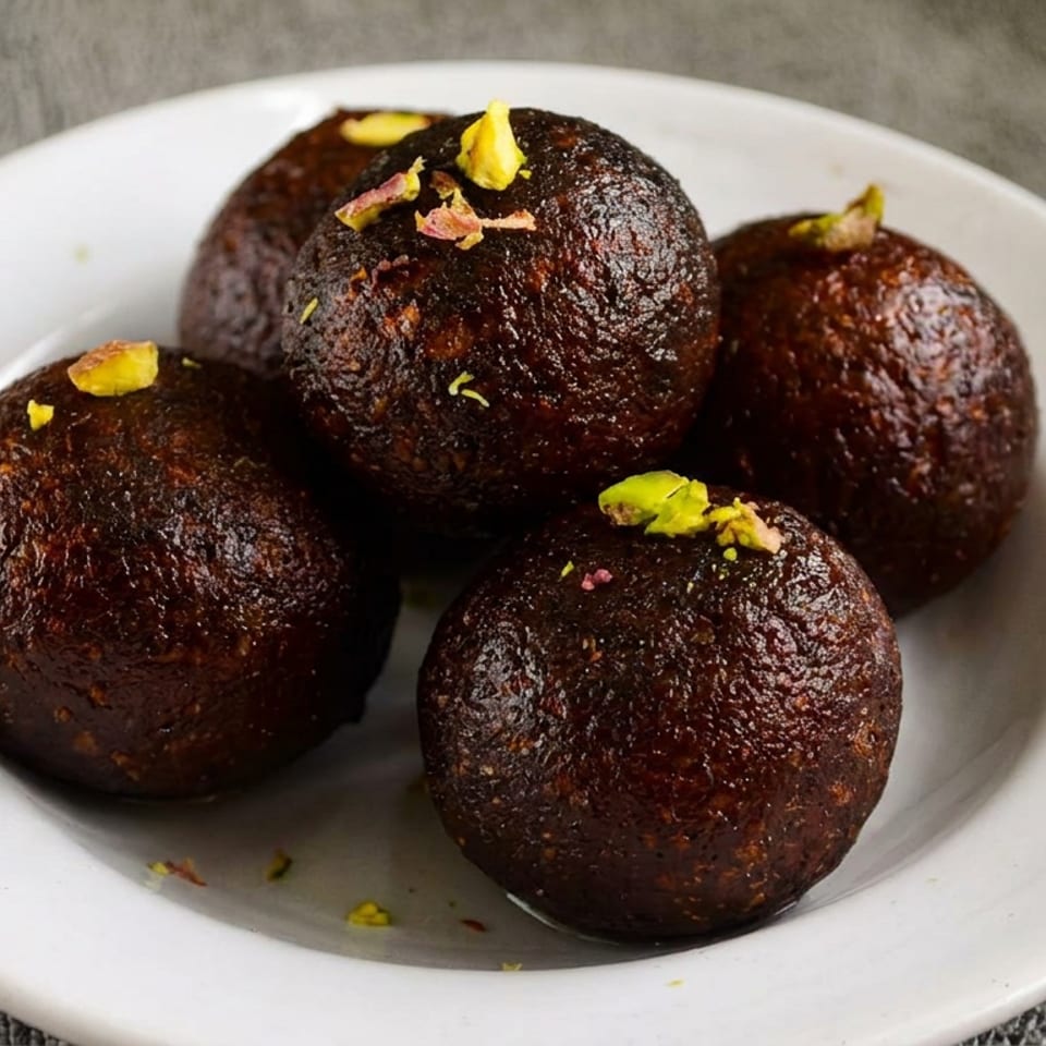 Four dark brown round sweets garnished with pistachios on a white plate.
