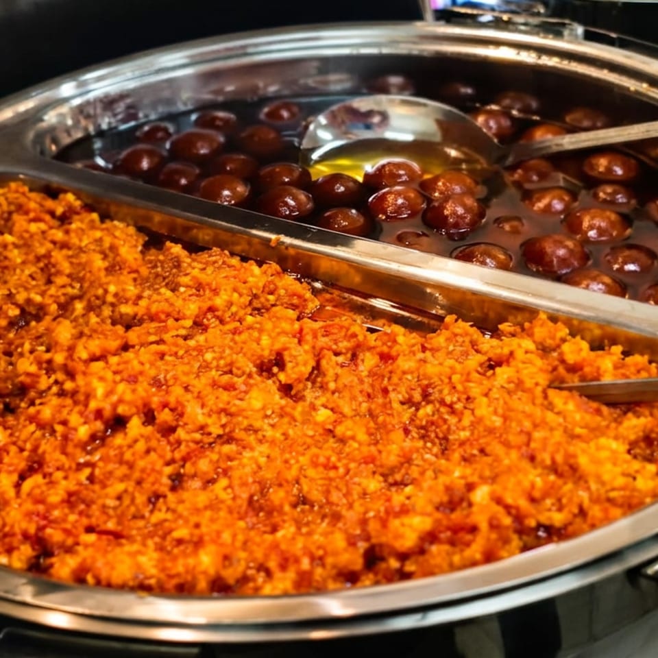 Stainless-steel buffet trays hold orange spiced rice and round brown sweets in syrup, with serving spoons.